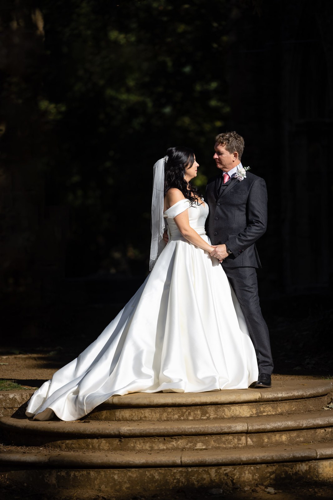 The couple face each other on curved stone steps in dappled light