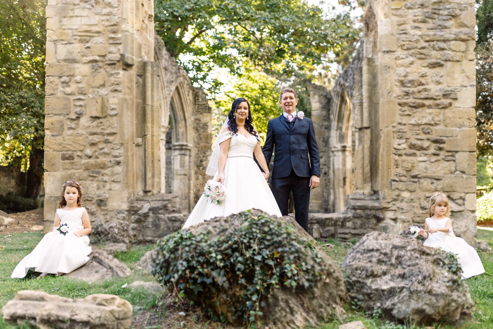 The couple stand among the abbey ruins with the flower girls seated on rocks
