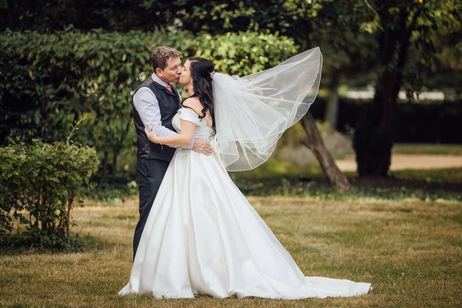 The veil lifts on the breeze as the couple kiss on the lawn by the hedges