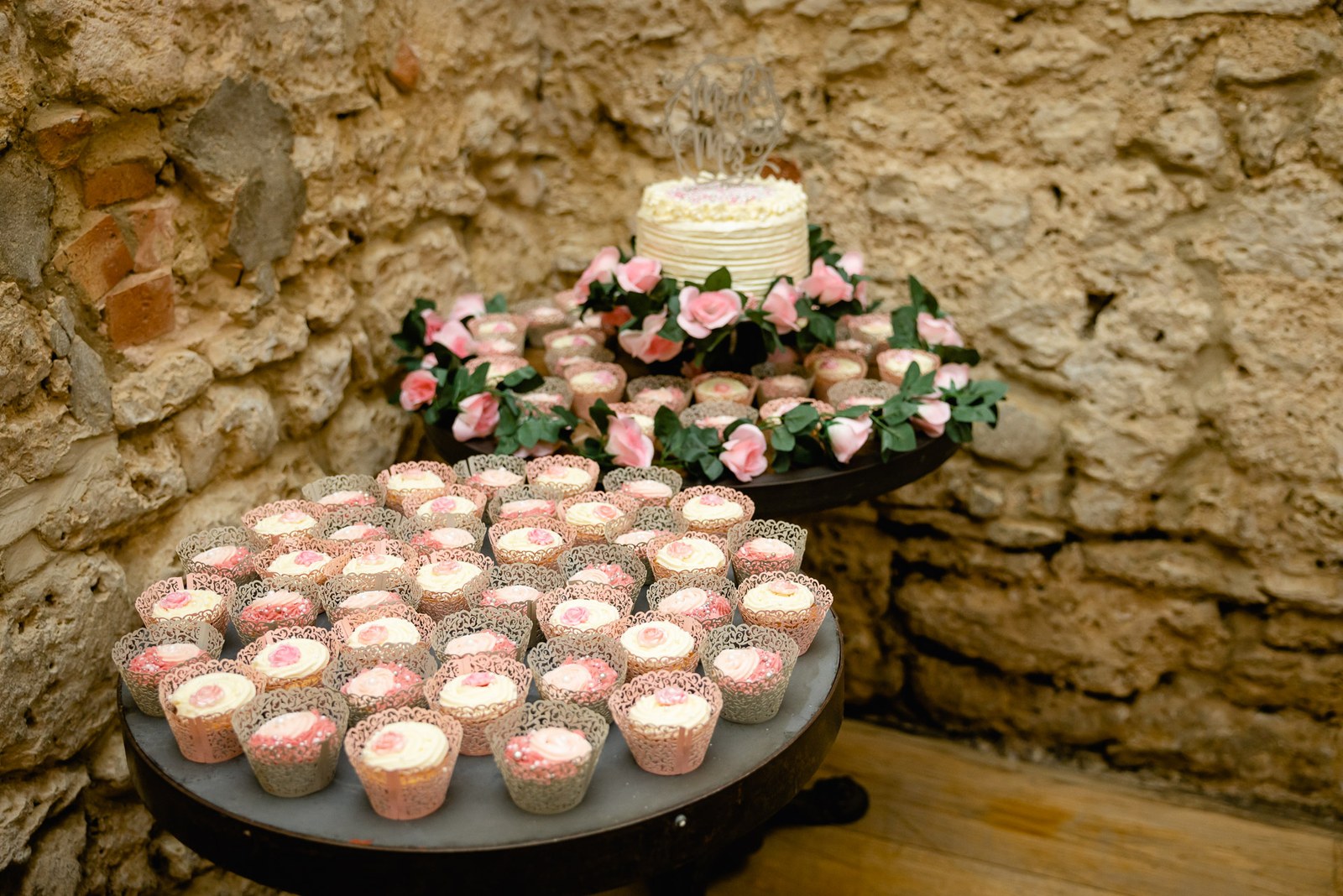 A dessert table of cake and iced cupcakes with pink roses against the stone wall