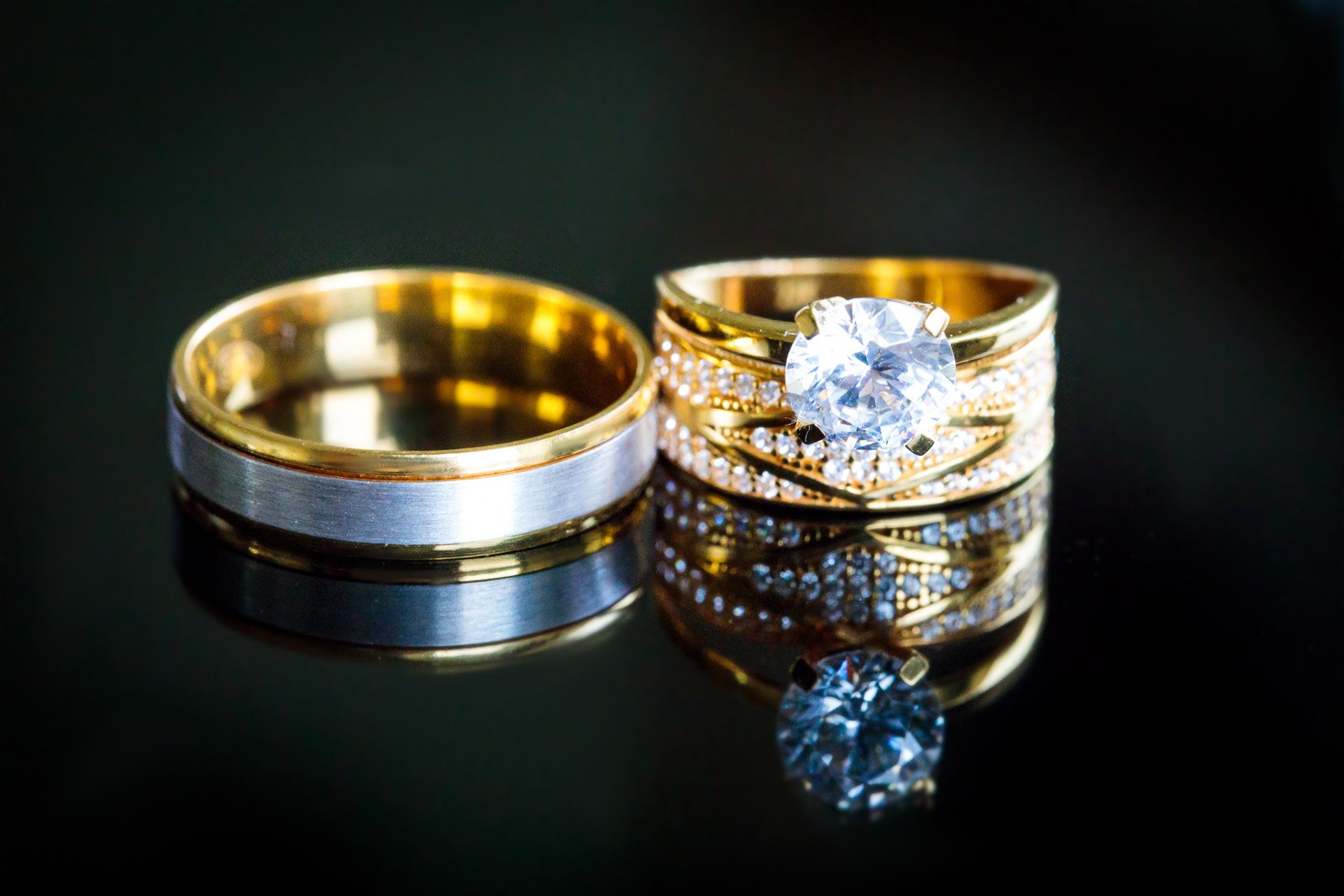 A two-tone gold band and a diamond solitaire ring reflected on a black surface