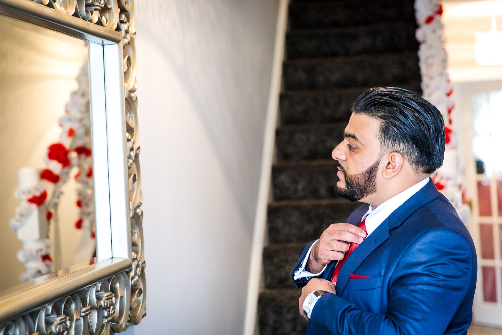 The groom adjusts his red tie before a mirror on the staircase