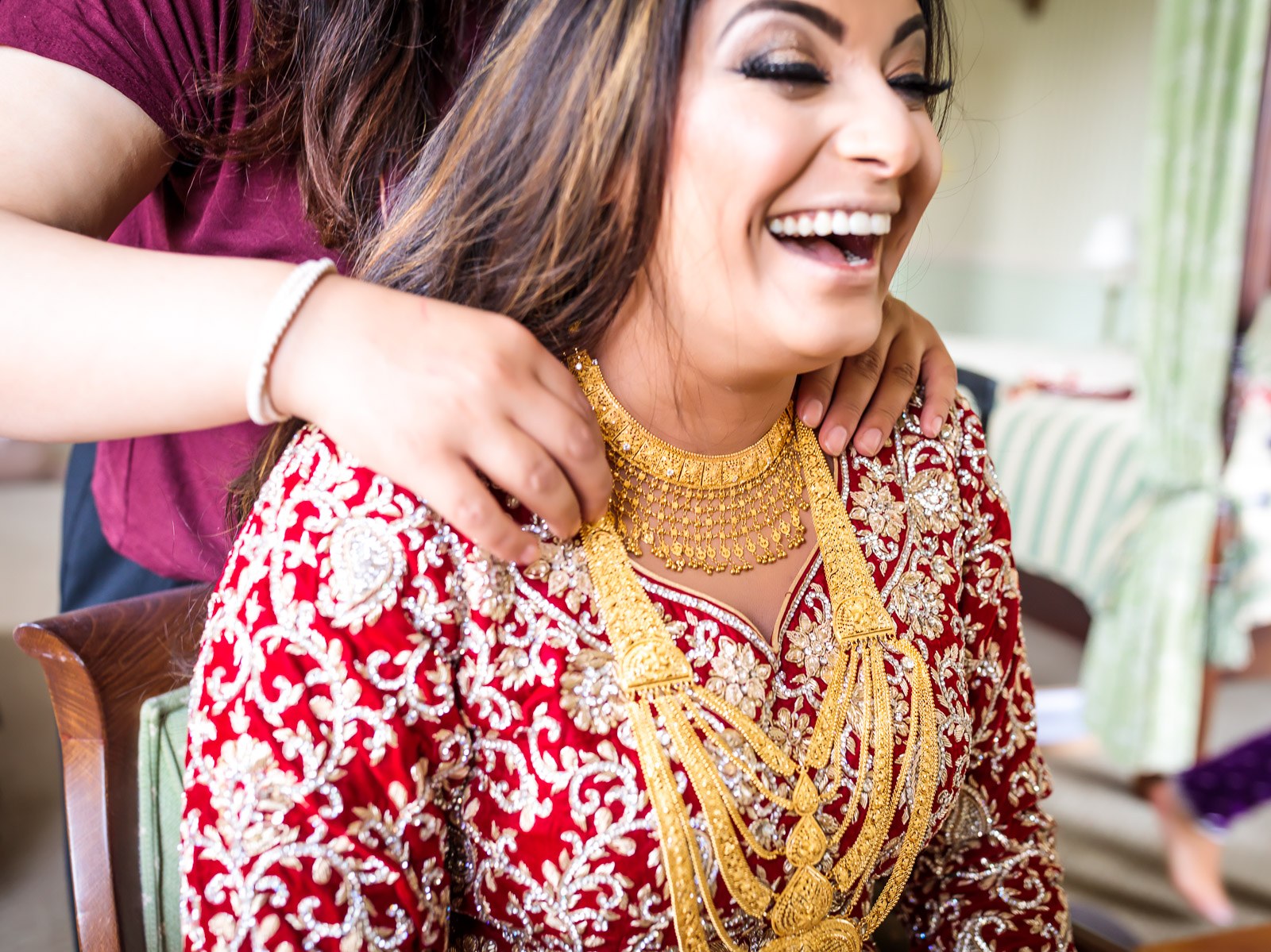 The bride laughs as a helper fastens her gold necklace over red bridal wear