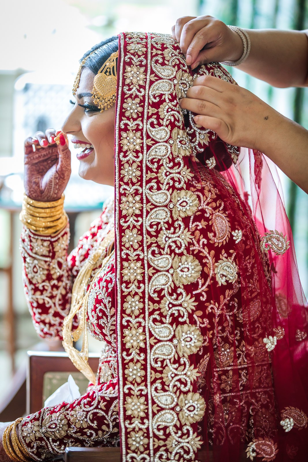 A helper drapes the bride's red dupatta as she smiles