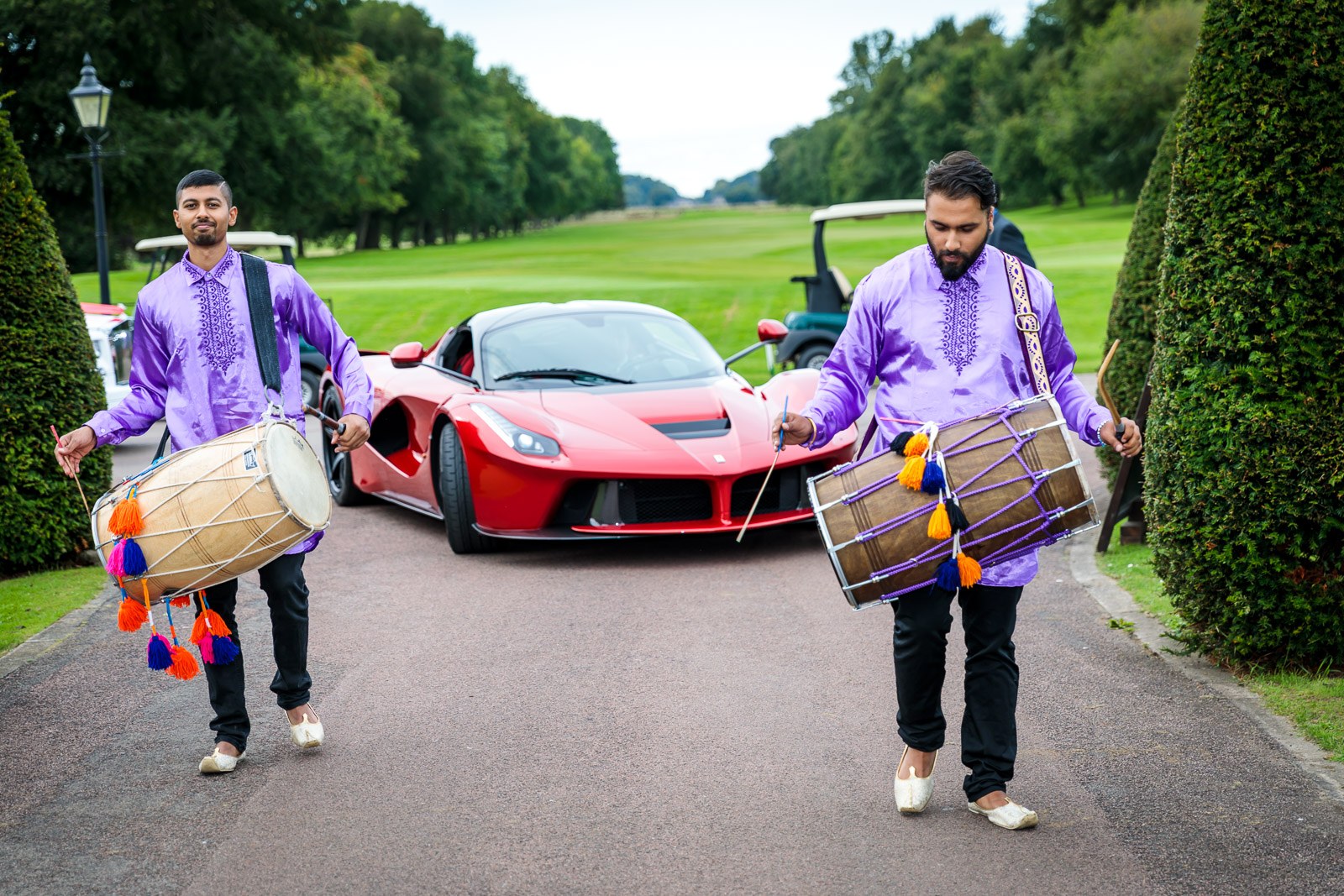 Two dhol drummers in purple flank a red Ferrari on the driveway