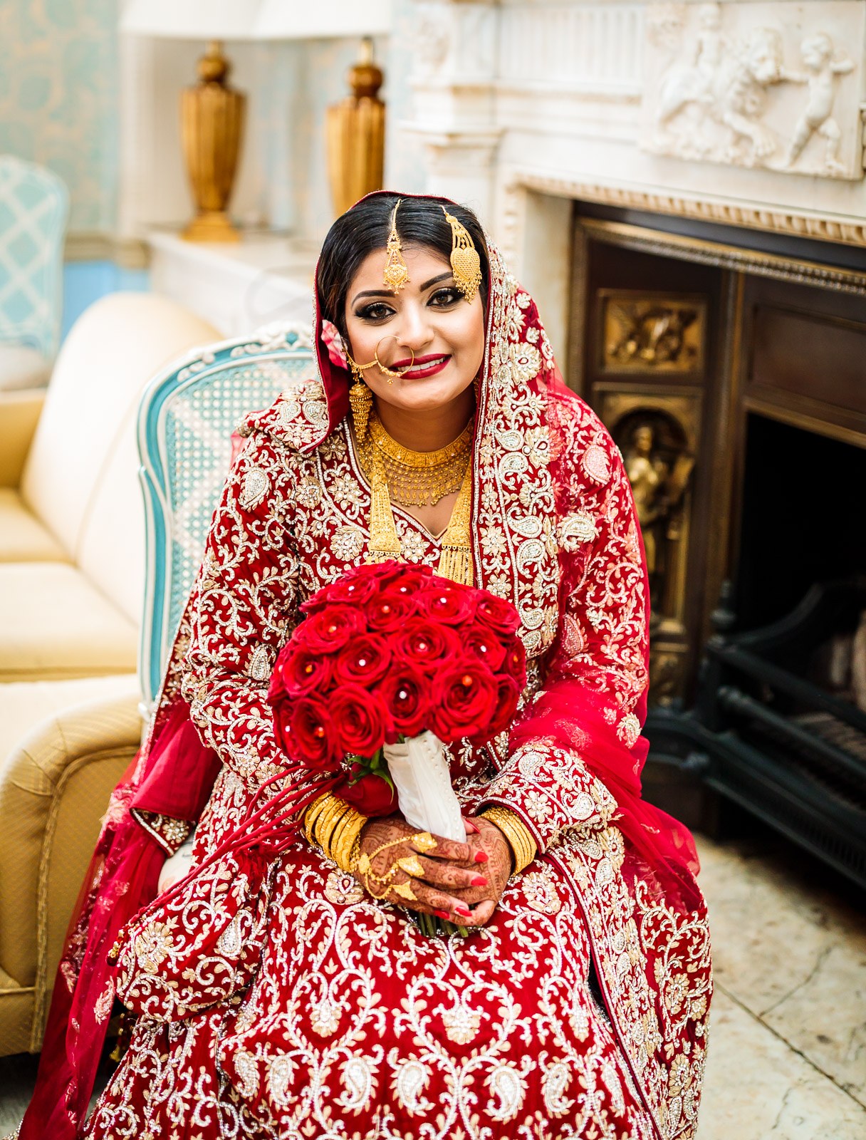 The bride sits in red bridal wear with a red rose bouquet by an ornate fireplace
