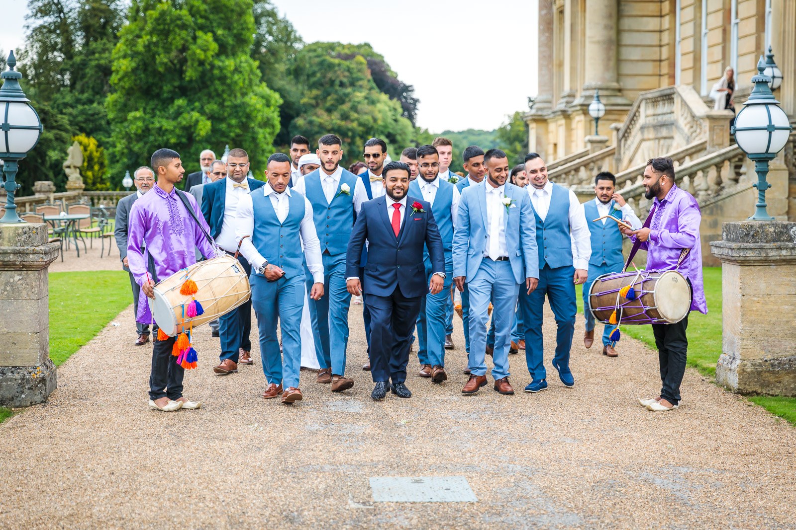 The groom and groomsmen walk with a dhol drummer outside Heythrop Park