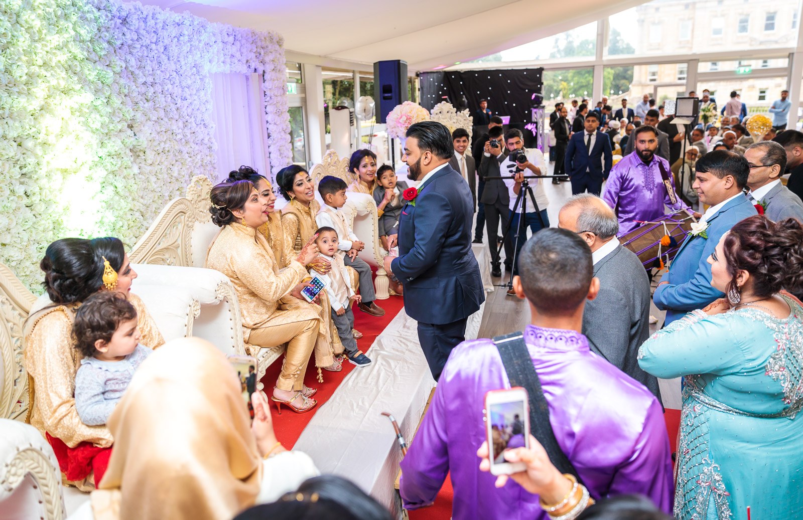 Guests fill the glass marquee before a white flower-wall stage