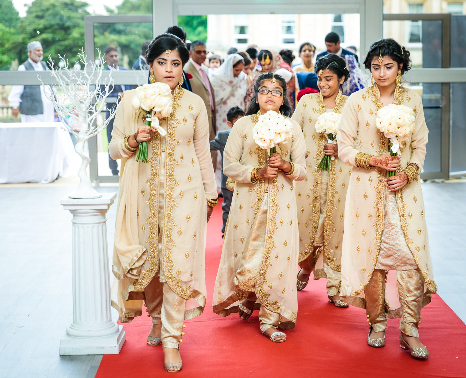Bridesmaids in cream carry white bouquets down the red carpet into the marquee
