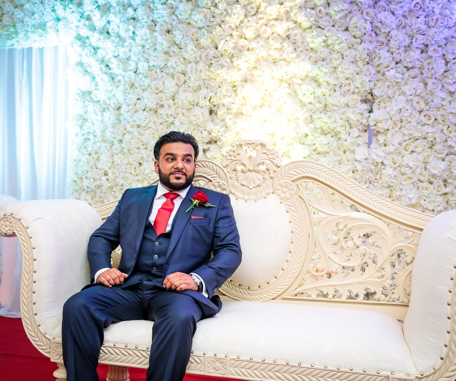 The groom sits alone on the ornate stage sofa before the flower wall