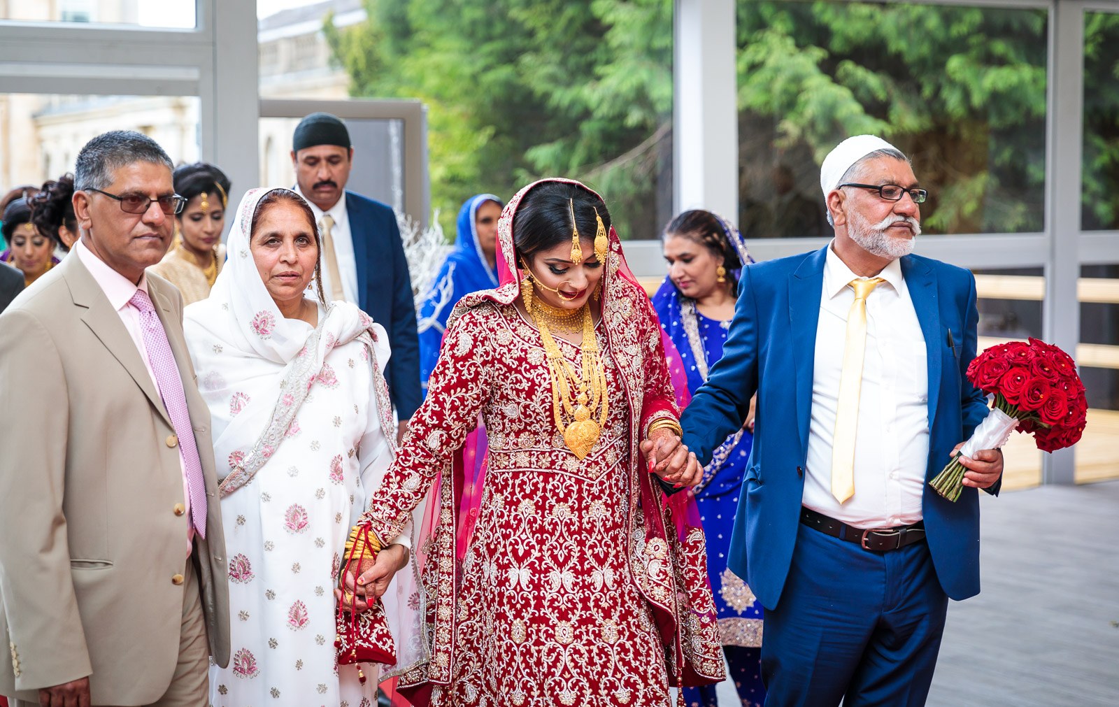 The bride is escorted in by her parents, one carrying her red rose bouquet