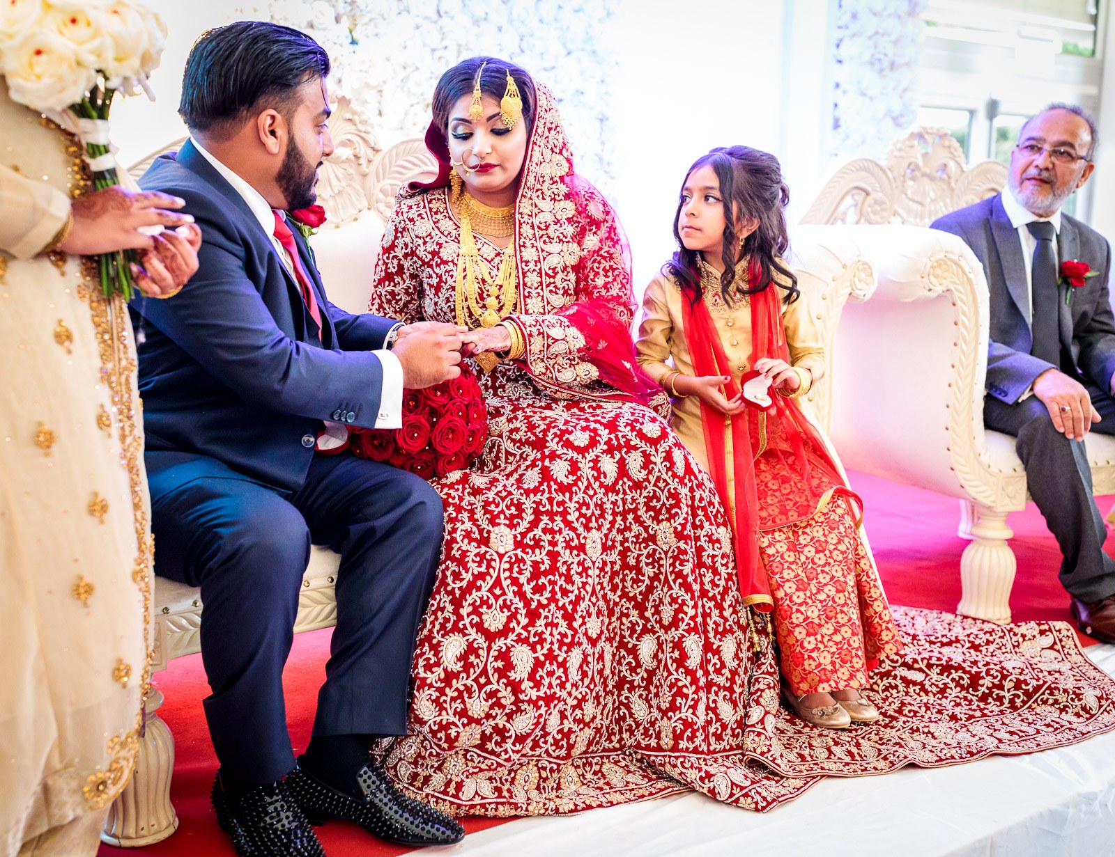 The groom slides a ring onto the bride's hand on the stage sofa