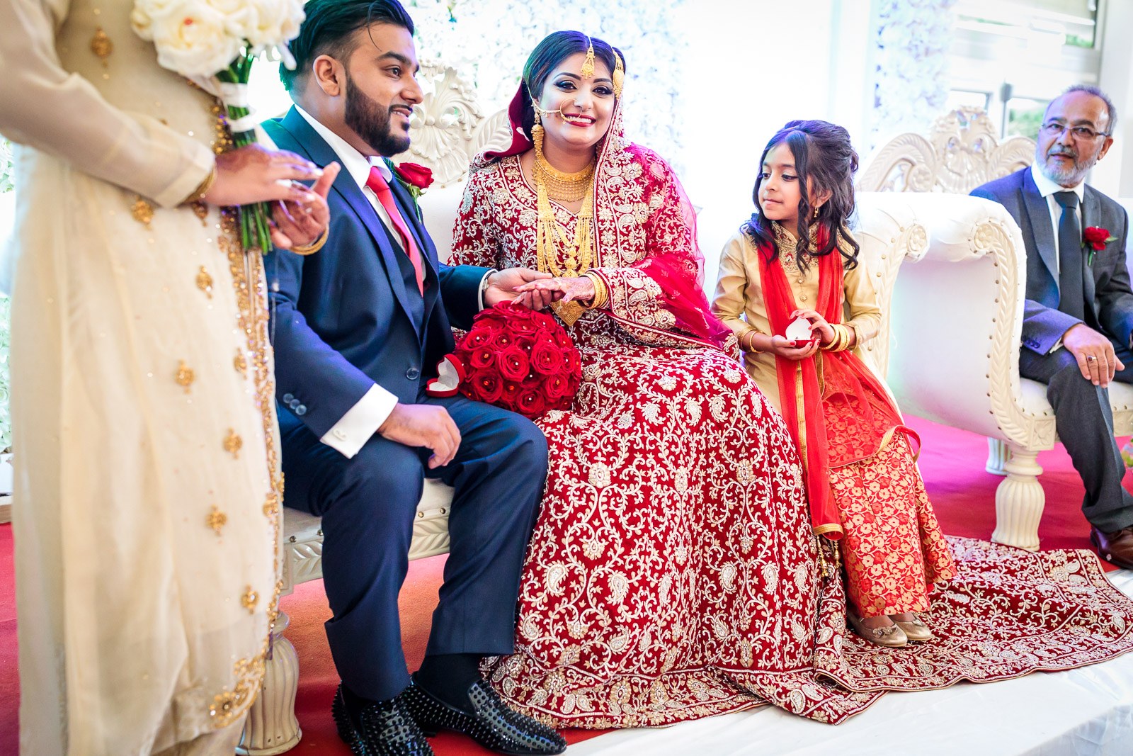 The couple sit on the stage, the bride's red bouquet resting between them