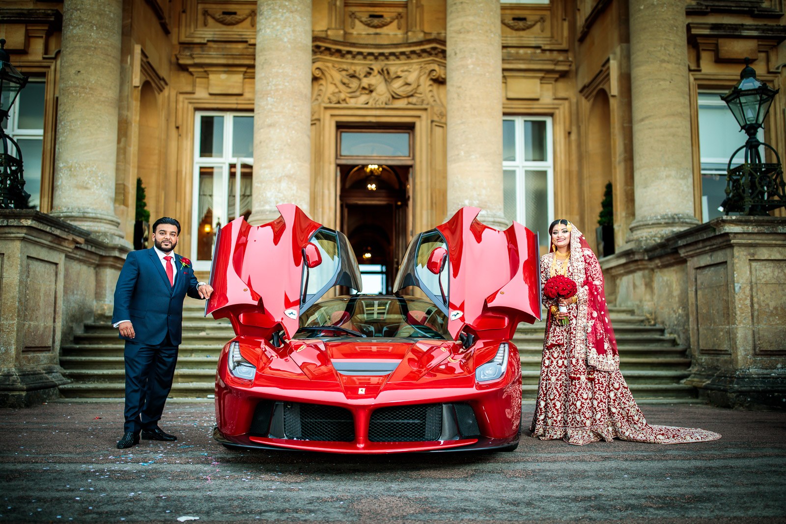 The couple stand either side of the red Ferrari, doors raised, before the mansion
