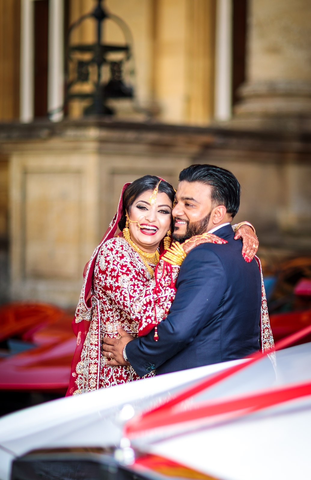 The couple embrace and laugh between the wedding cars at Heythrop Park