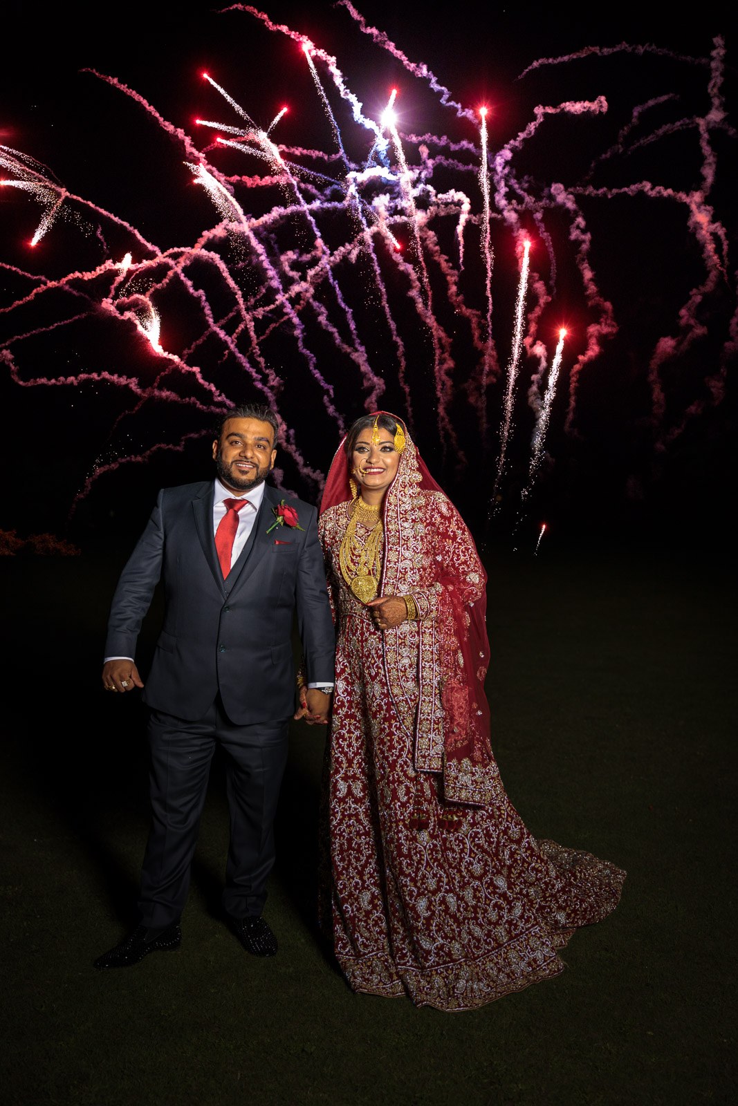 The couple stand on the lawn as pink fireworks light the night sky