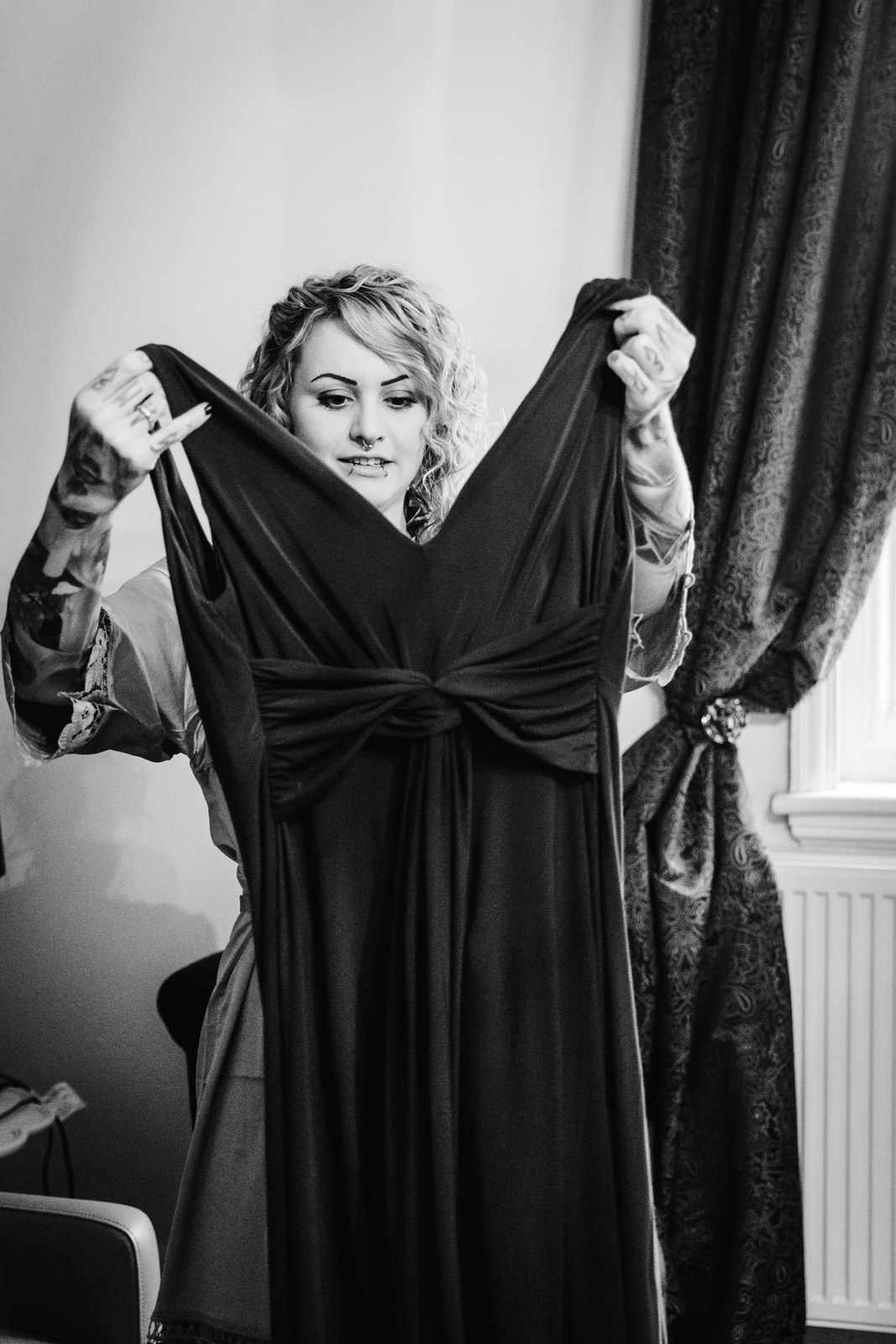 Black-and-white shot of a bridesmaid holding up her dark gown by the window