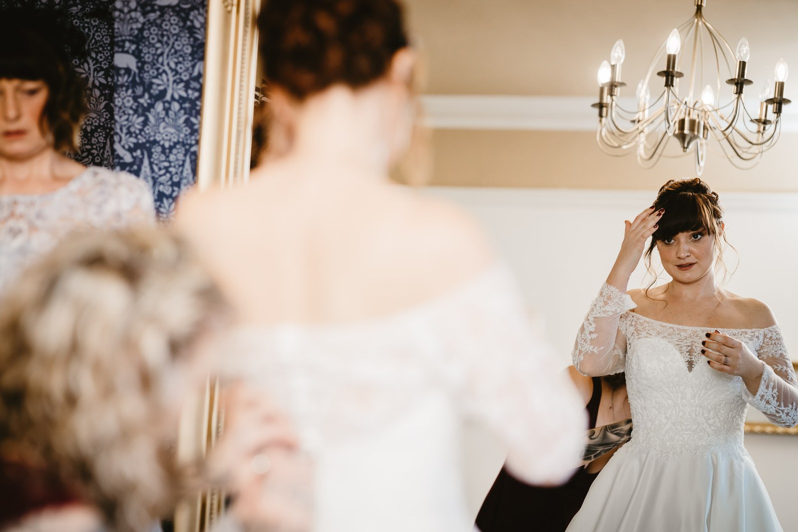 The bride is helped with her gown beneath a chandelier by the mirror