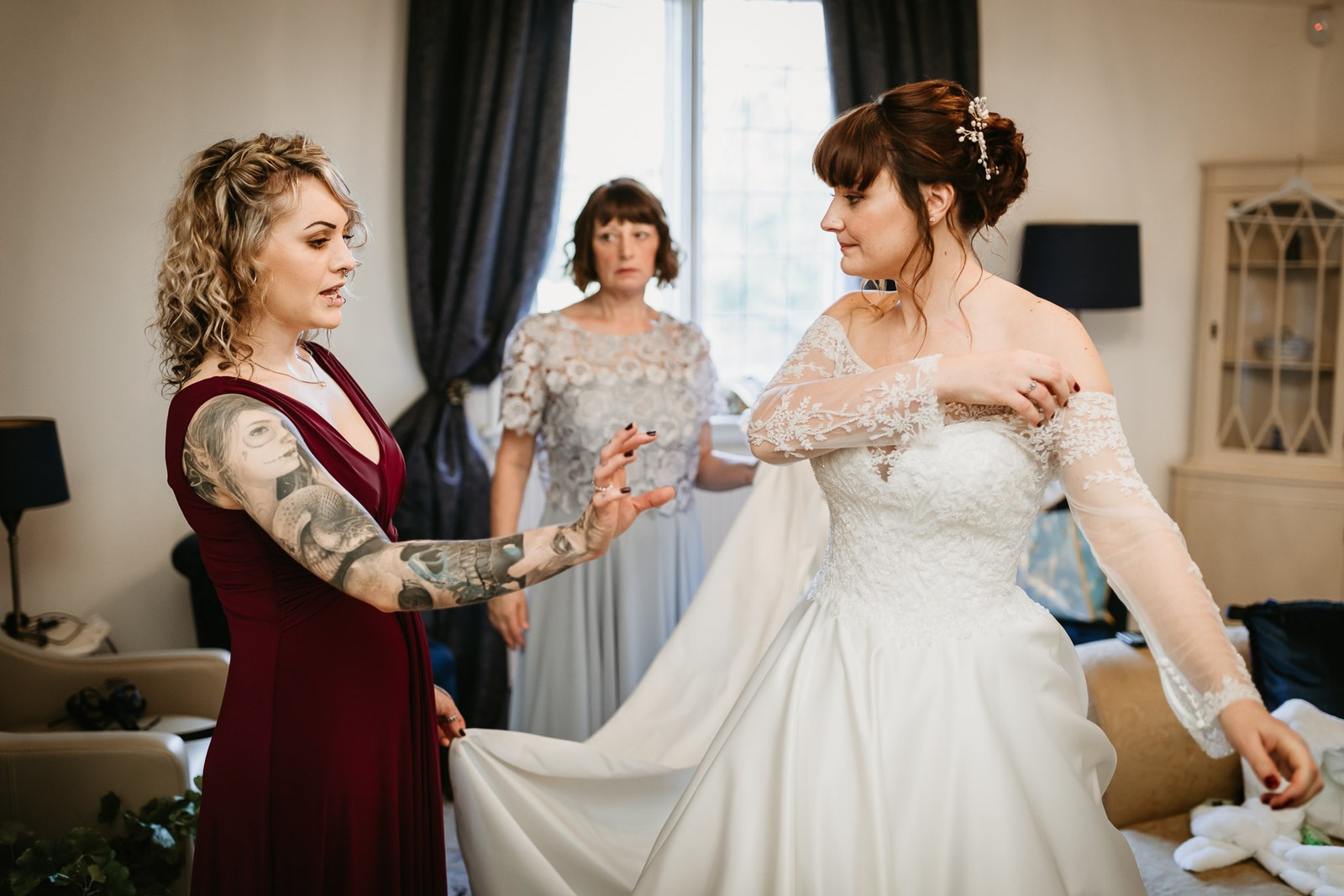 A bridesmaid in burgundy arranges the bride's gown at the window