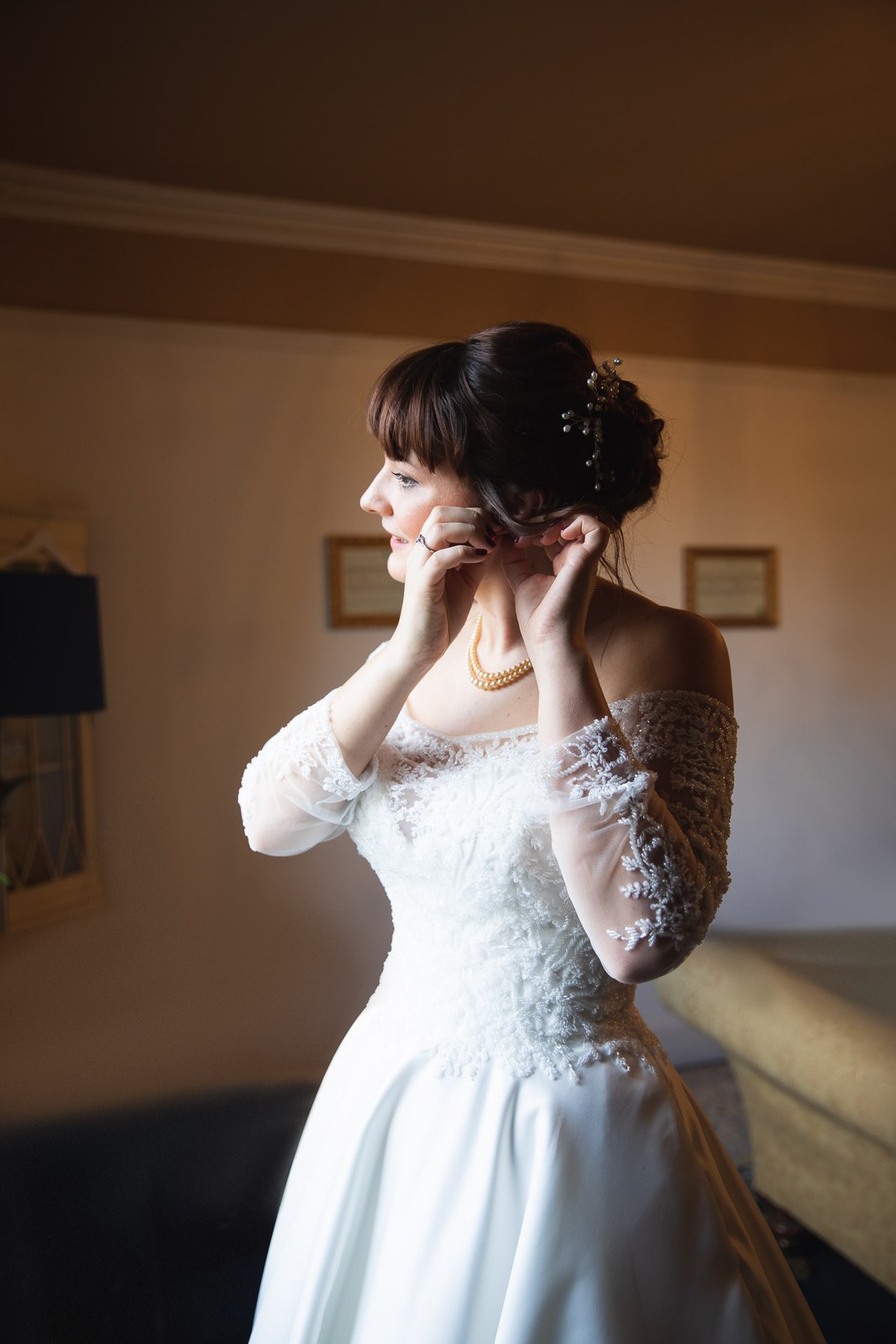 The bride fixes her earring by the window, pearls at her neck