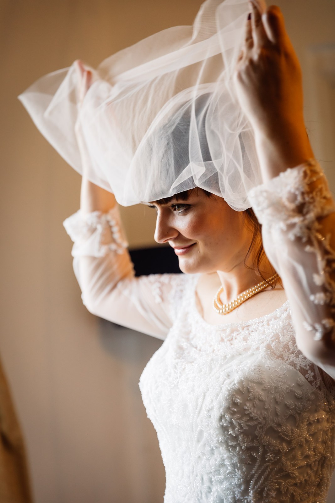 The bride lifts her veil in profile by the window