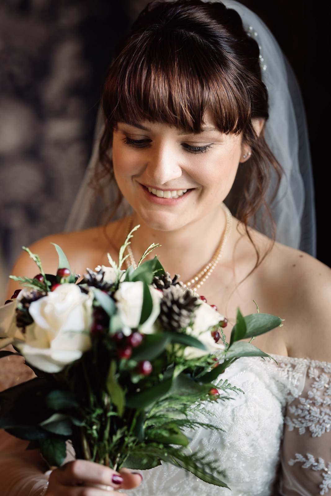 The bride smiles down at her winter bouquet of white roses and pinecones