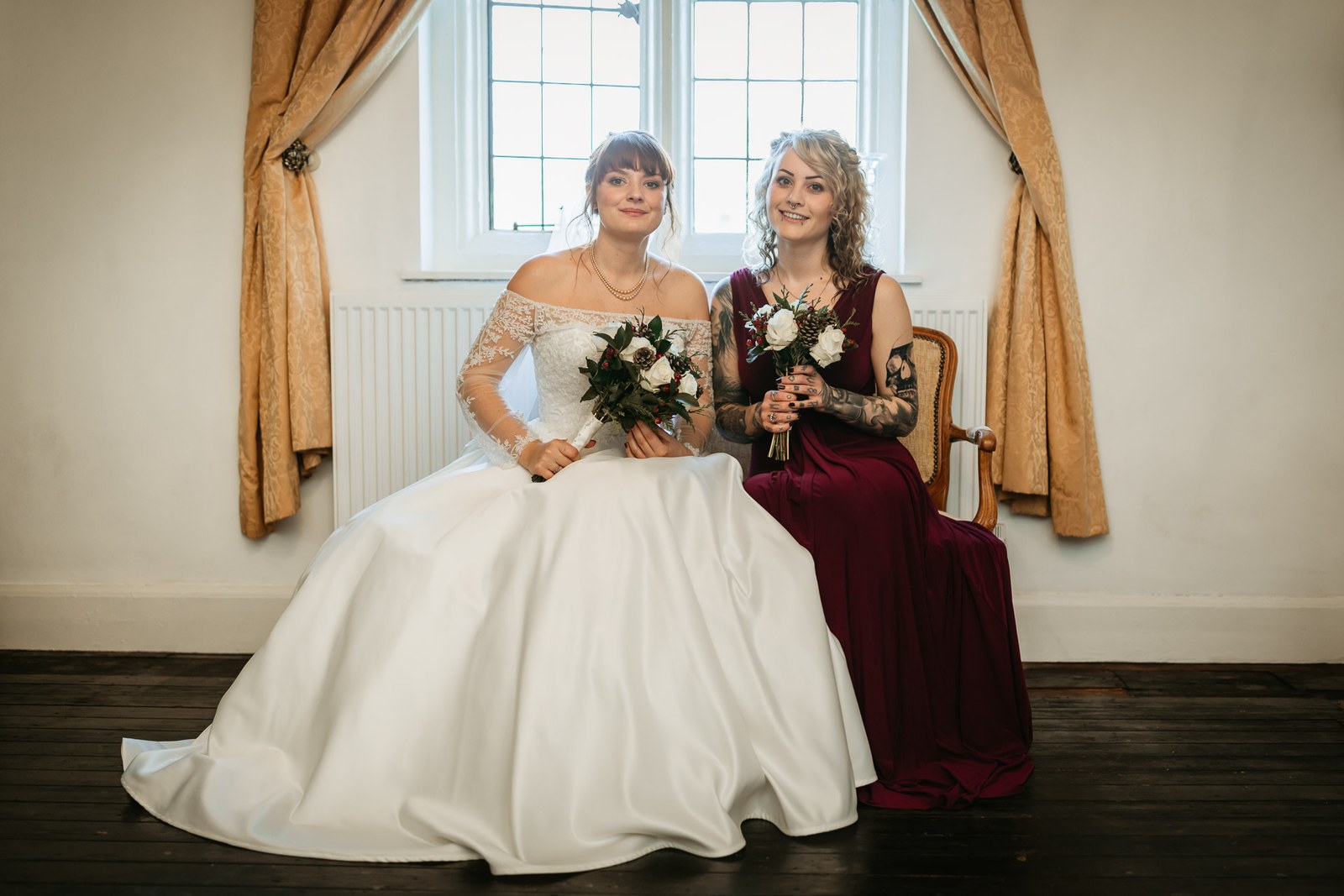 The bride and a bridesmaid in burgundy sit with their bouquets by the window