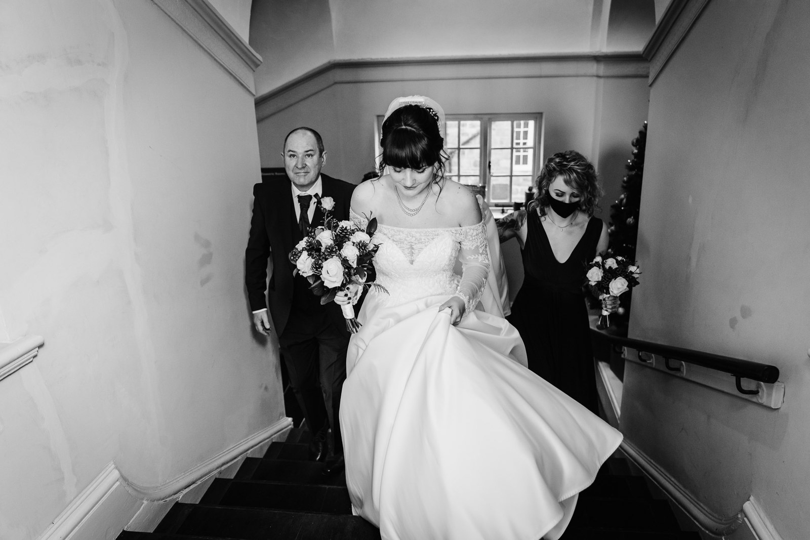 Black-and-white shot of the bride coming downstairs with her father and bridesmaid