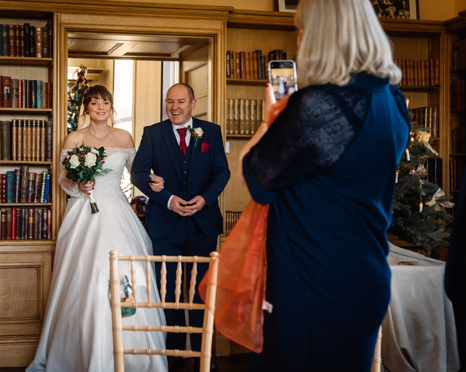 The bride enters the library on her father's arm as a guest takes a photo