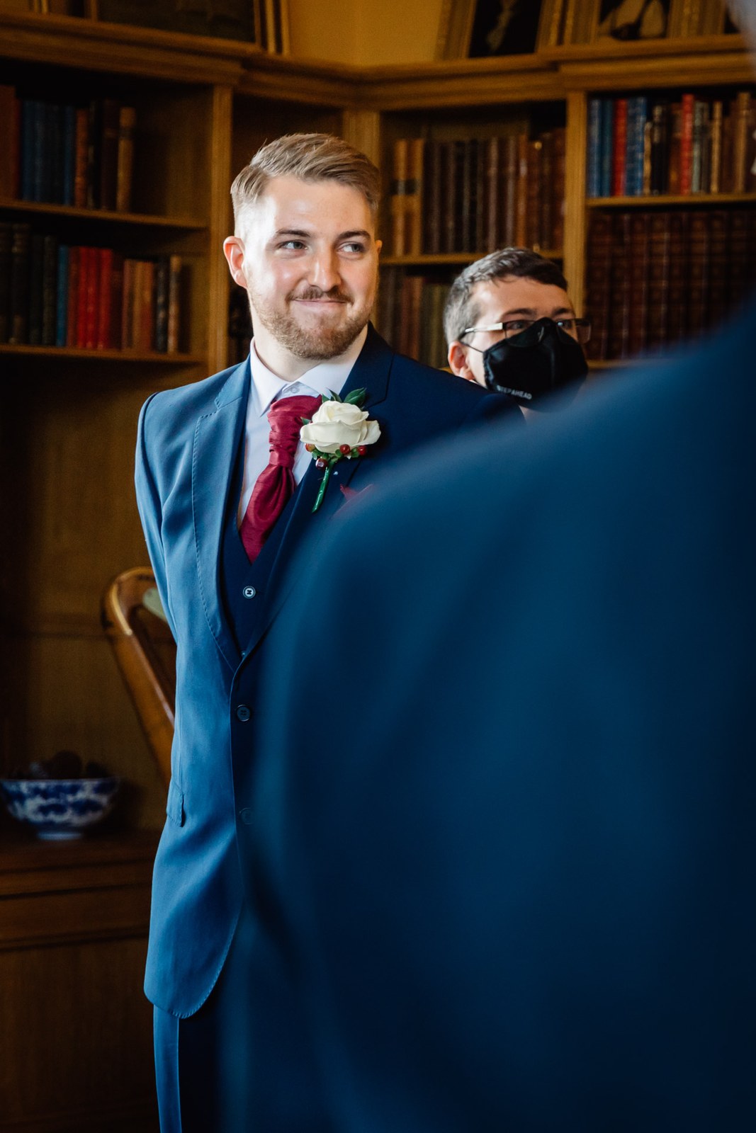 The groom waits in his navy suit before the library bookcases