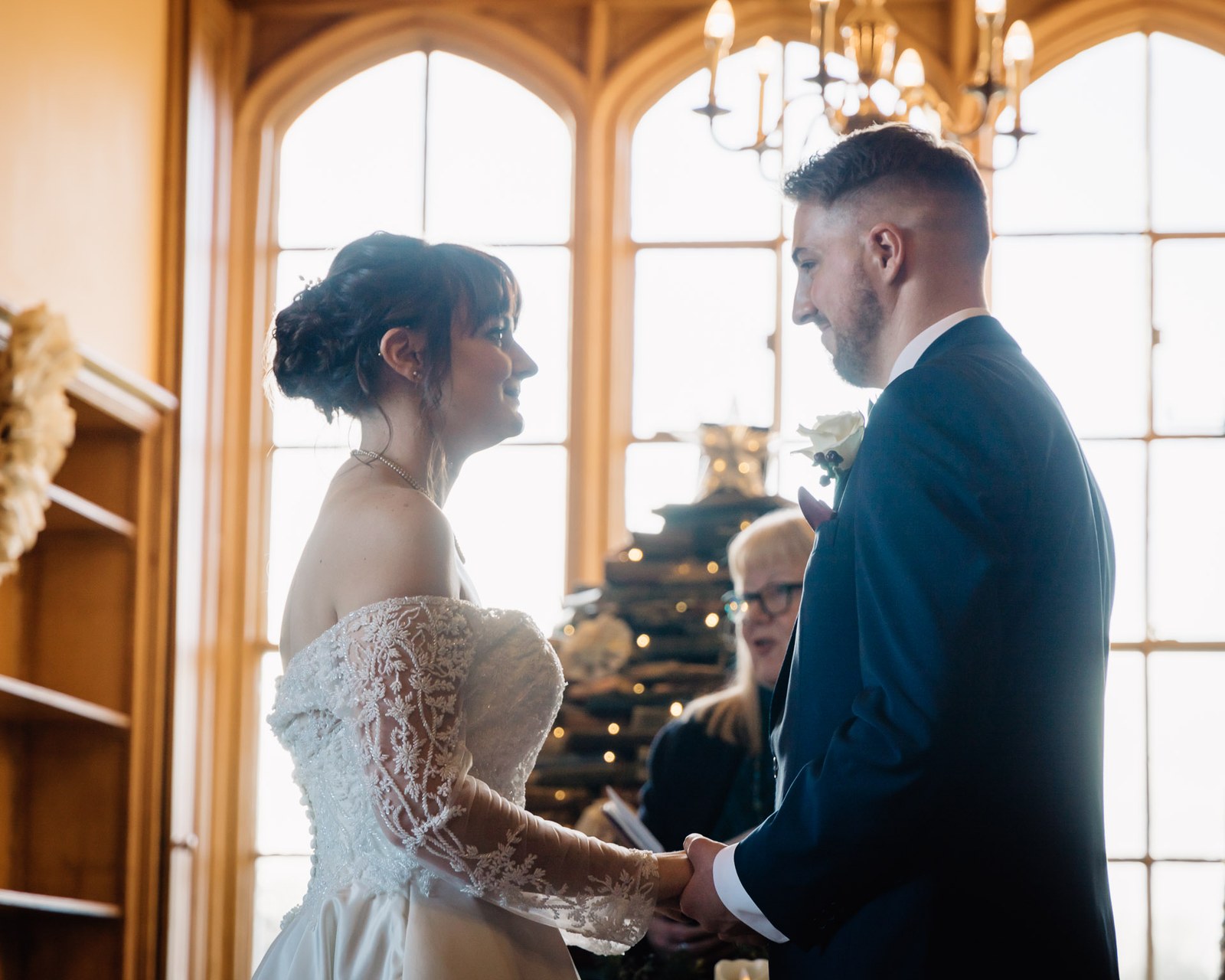The couple hold hands by the arched window, a Christmas tree behind