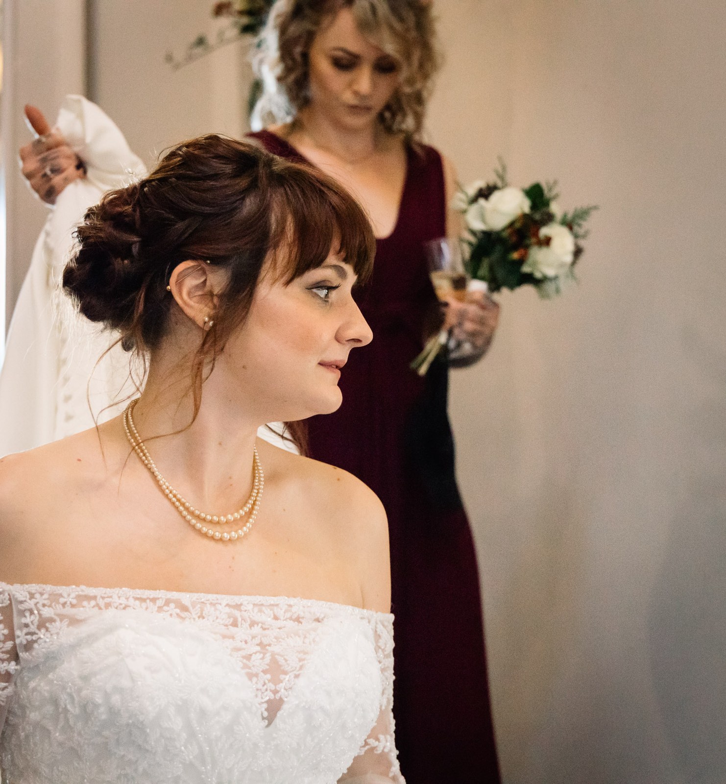 Portrait of the bride in profile, her bridesmaid in burgundy behind with a drink