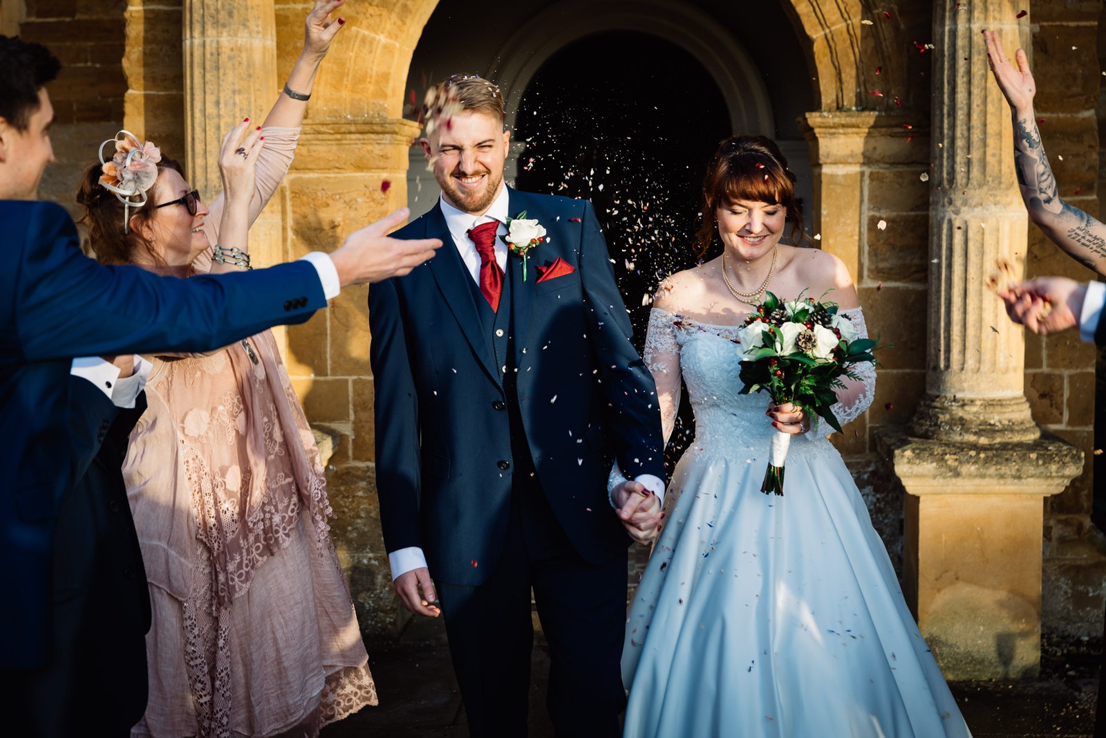 Confetti showers the couple beneath the golden-lit stone arch of Delapre Abbey
