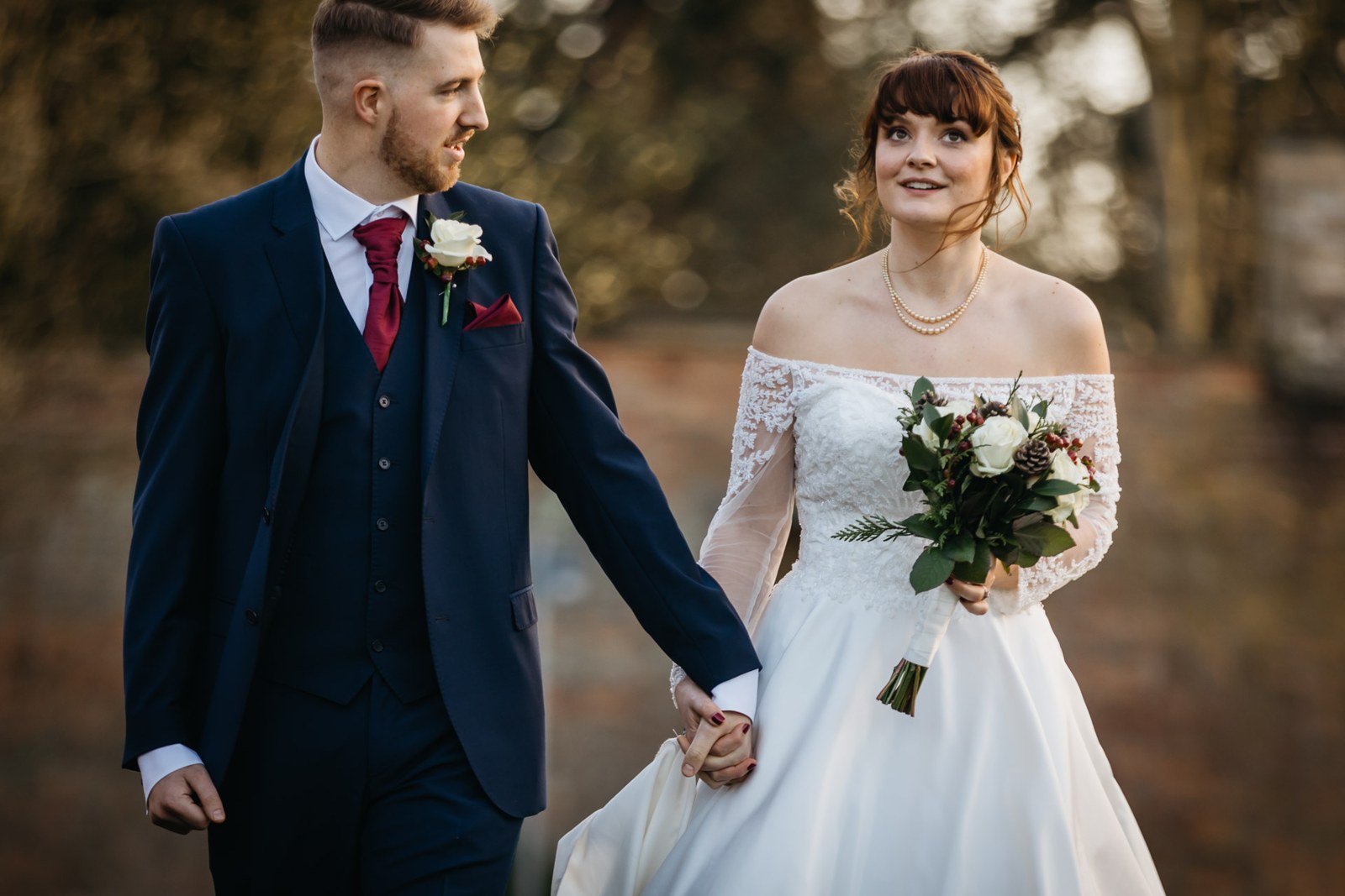Bride and groom walk hand in hand through the winter garden, her bouquet in hand