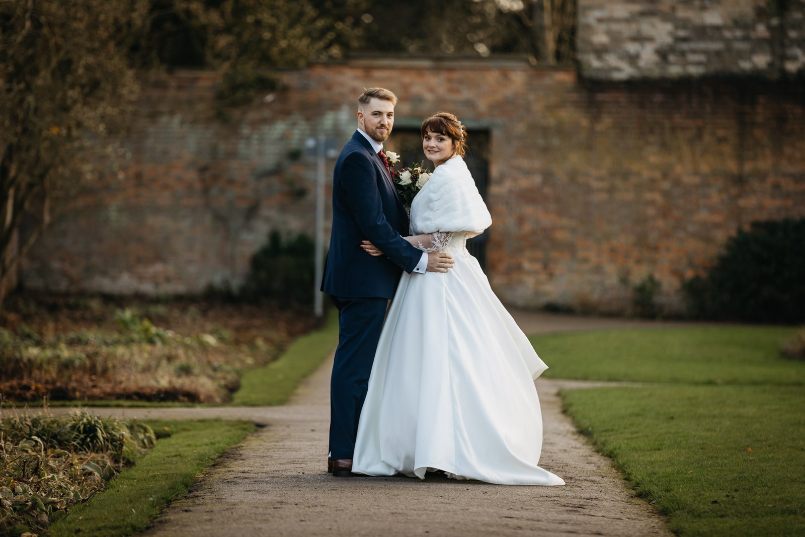 The couple embrace on a path by the old brick wall, the bride in a fur stole