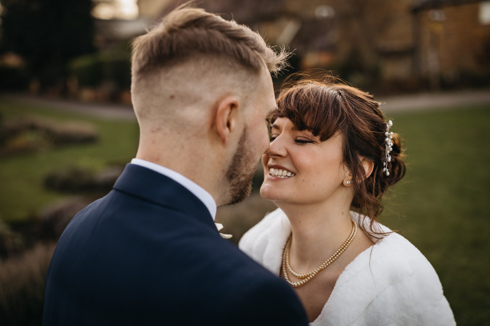 The bride in a fur stole and gold necklace stands close with the groom