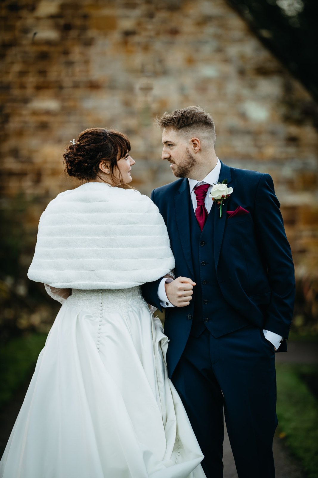 The couple face each other by a brick wall, the bride in her fur stole