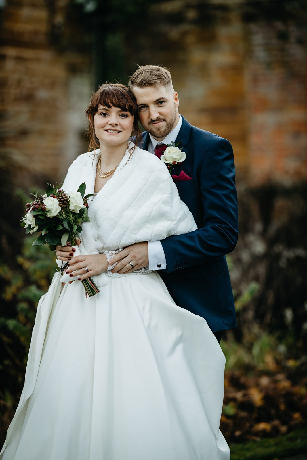 The couple embrace by the brick wall, the bride smiling with her bouquet