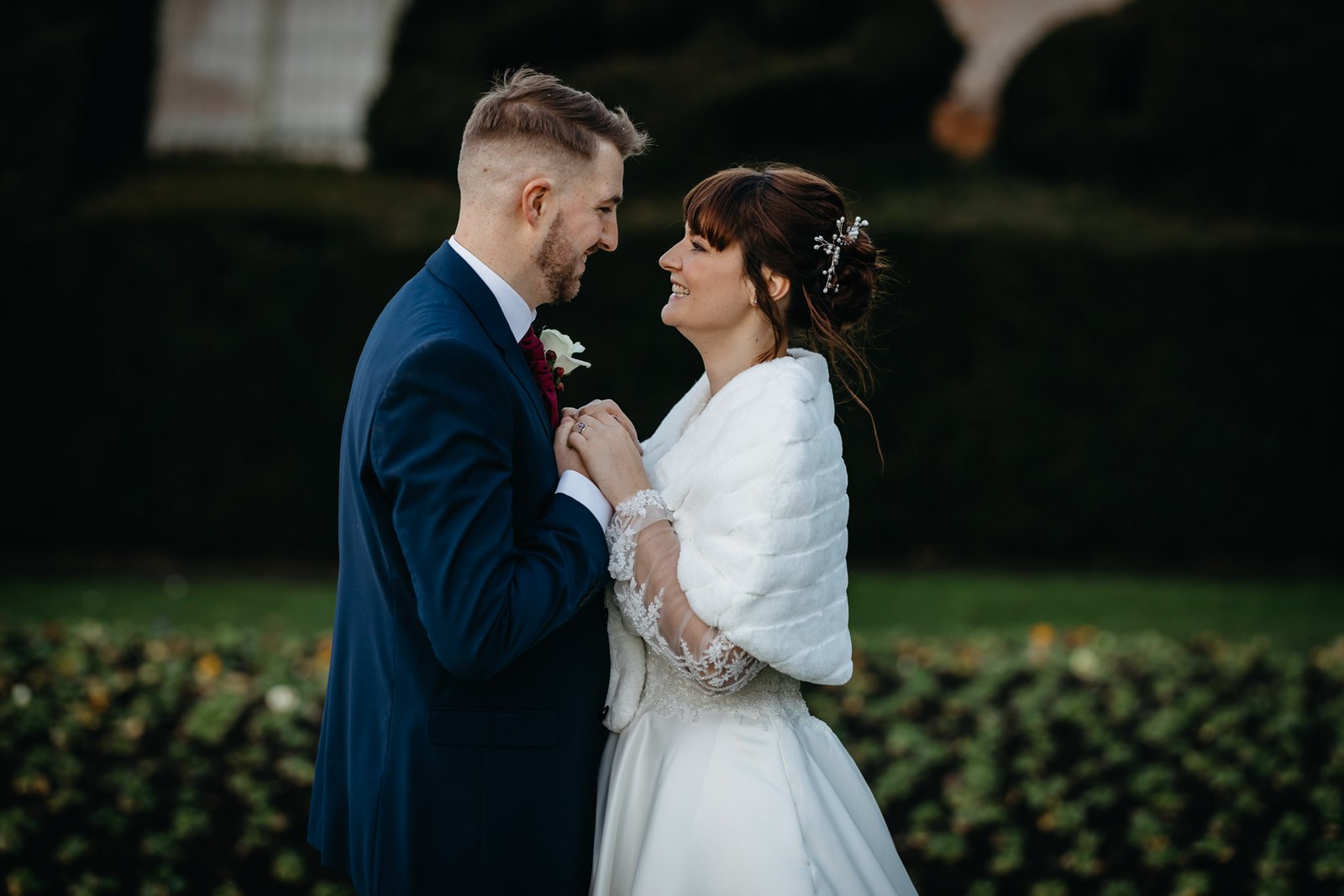 The couple lean nose to nose beside the yew hedges