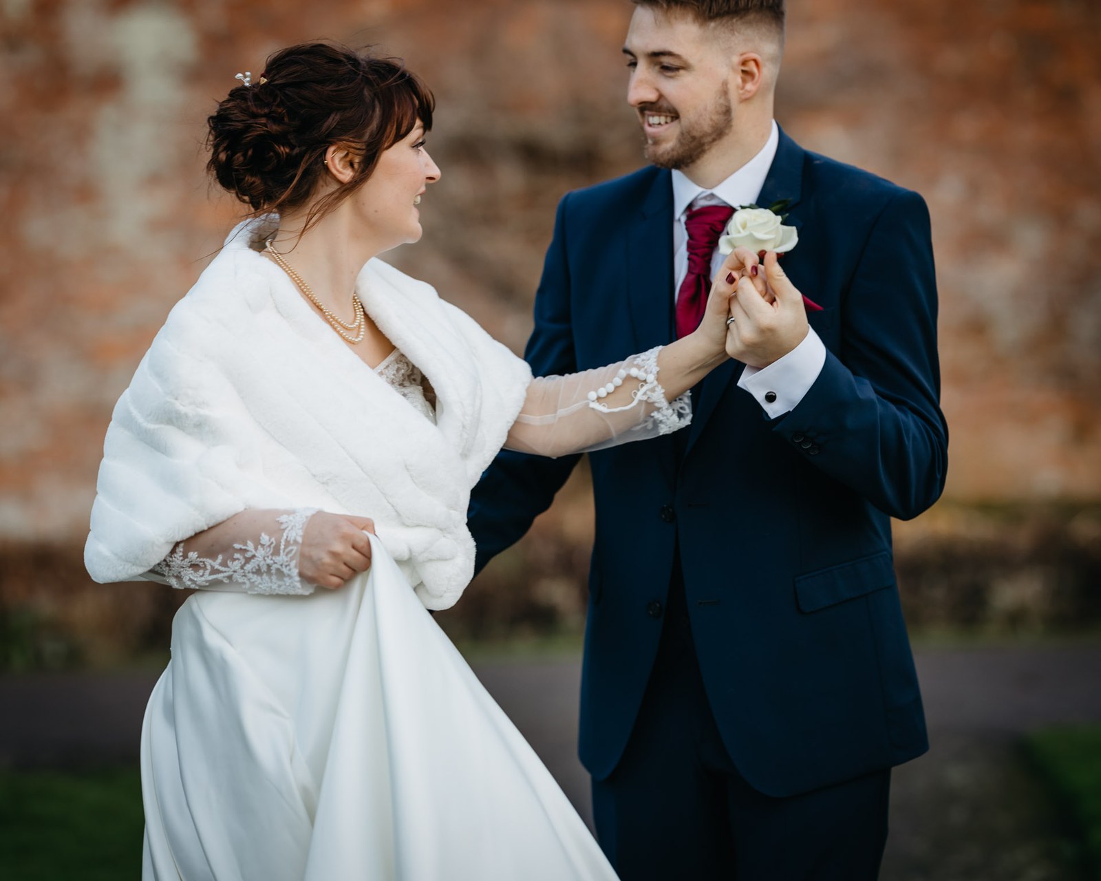 The groom lifts the bride's hand as she gathers her gown by the brick wall