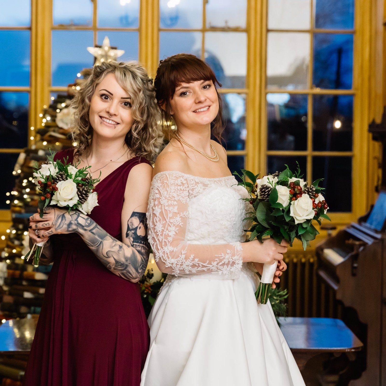 The bride and a bridesmaid in burgundy pose by the Christmas tree with their bouquets