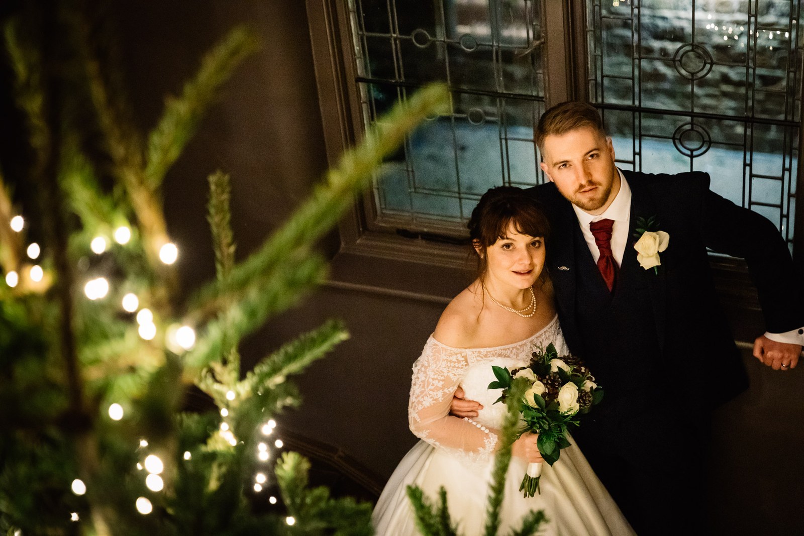 The couple stand by a fairy-lit Christmas tree, the bride holding her bouquet