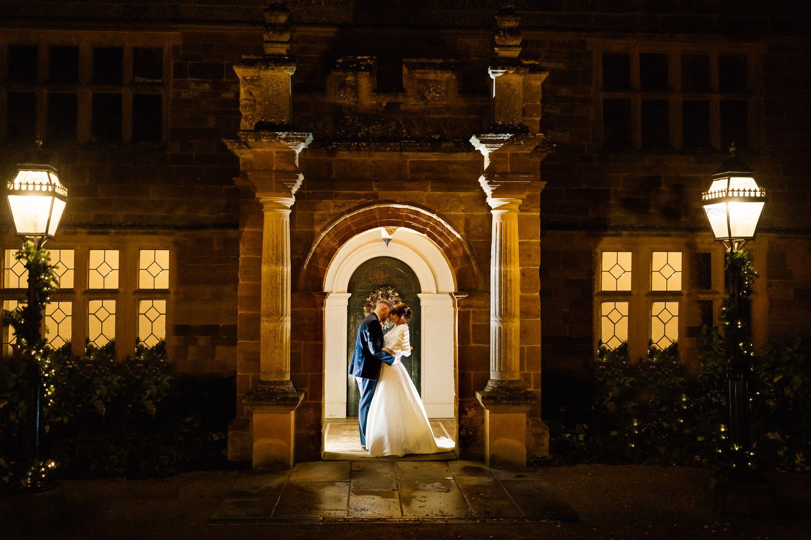 The couple embrace in the lamplit arched stone porch of Delapre Abbey at night