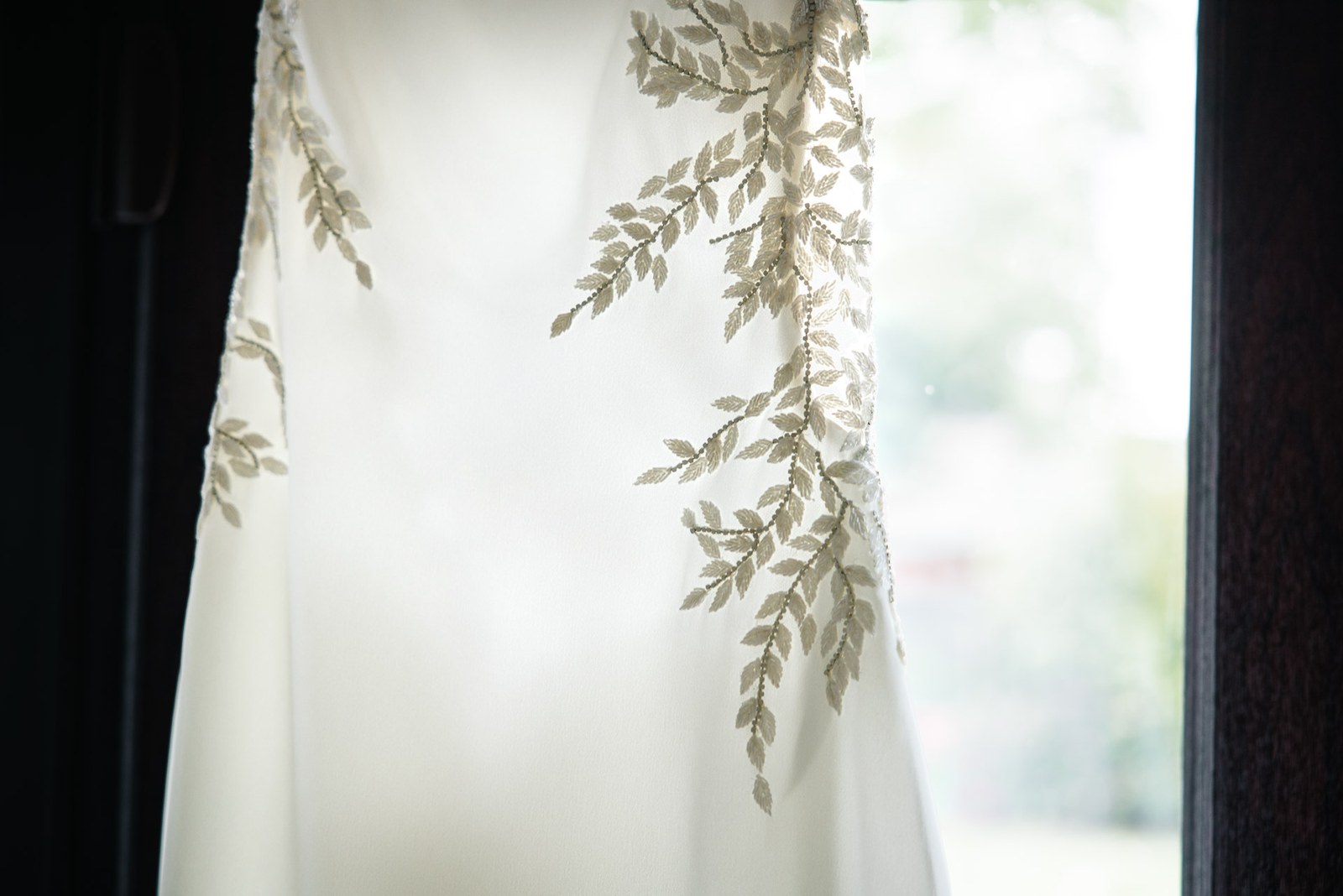 The bride's ivory wedding gown hanging by a window, its bodice trailing embroidered leaves