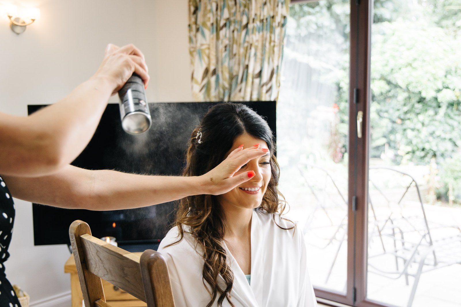 Stylist setting the bride's waves with hairspray beside a bright garden window