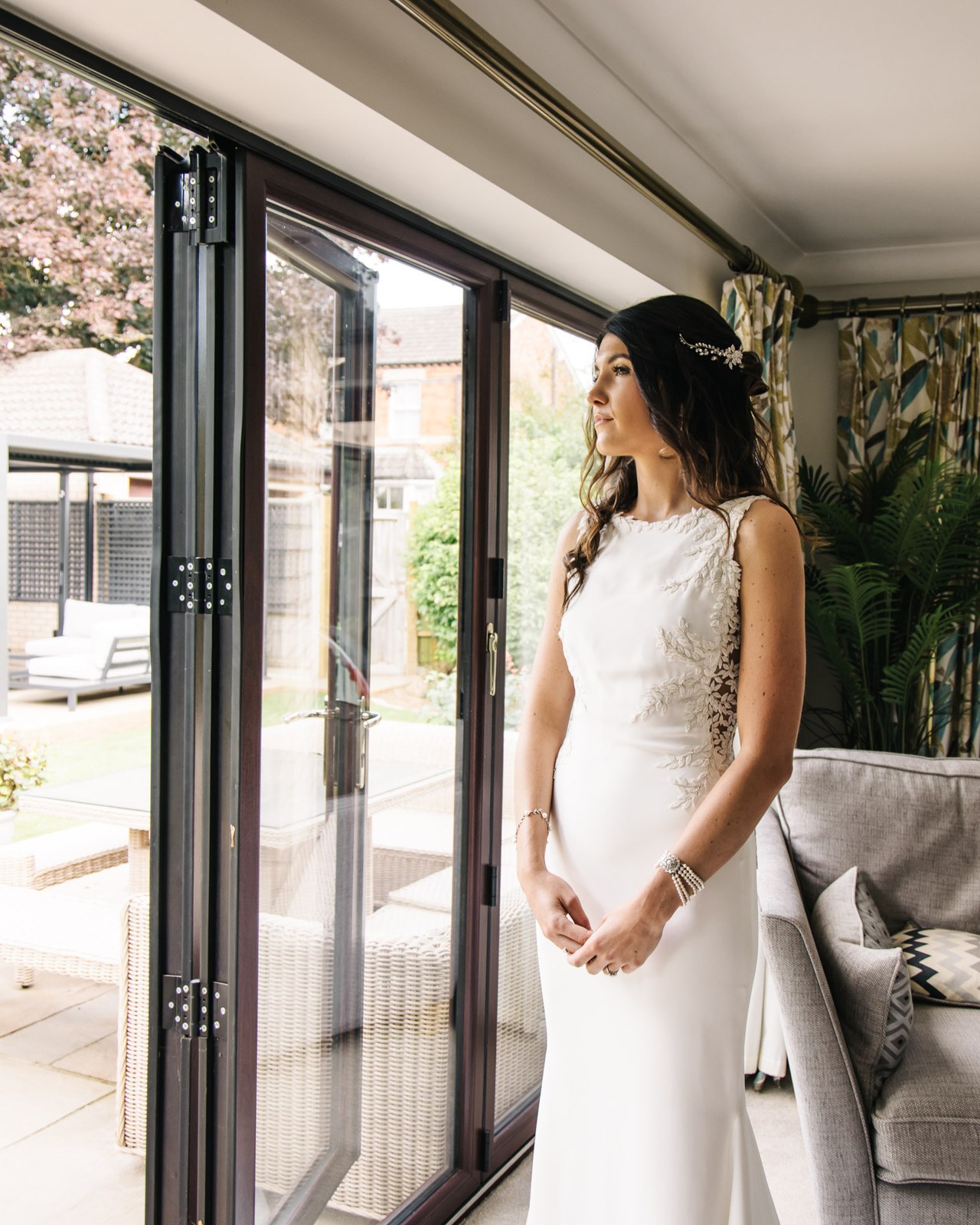 Bride in her fitted gown standing by open bifold doors, looking out to the garden