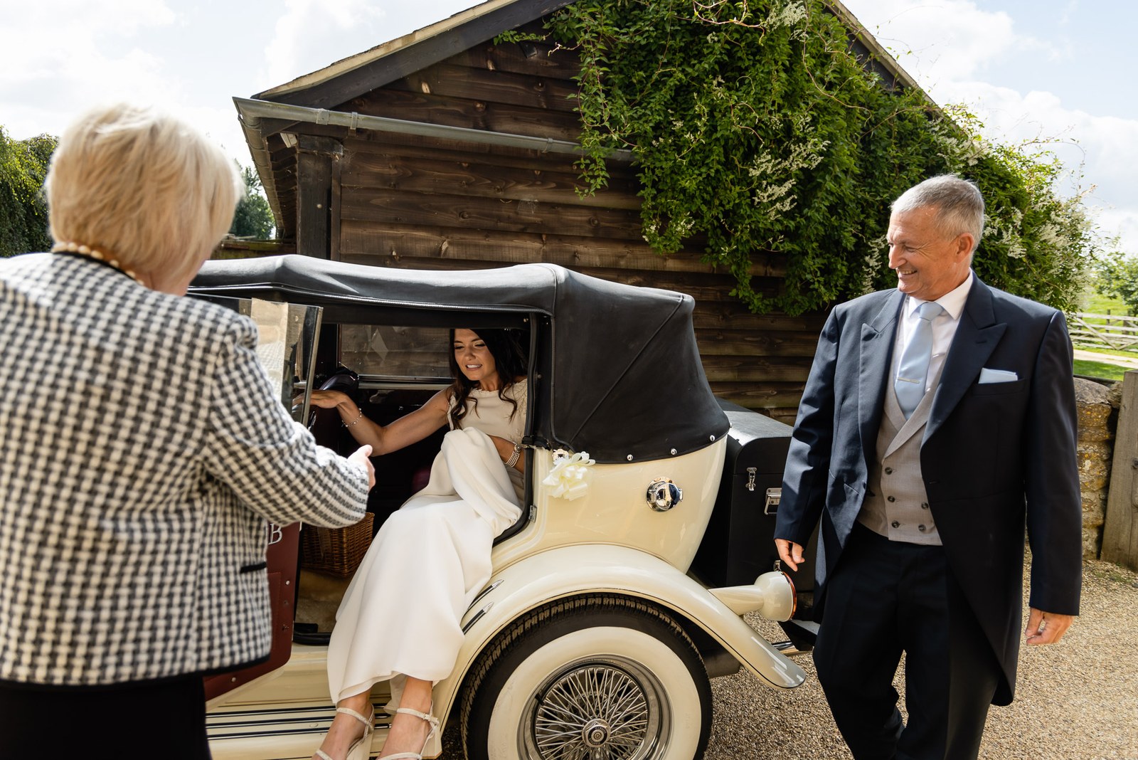 Bride arriving at Dodford Manor in a vintage cream car, helped from her seat by a waiting guest