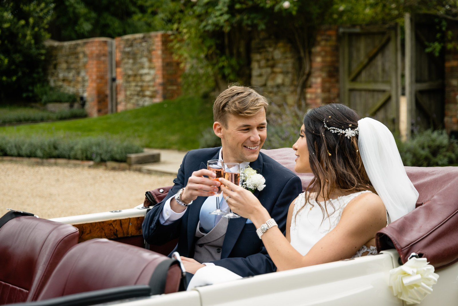 The couple toasting with drinks in an open-top vintage car in Dodford Manor's walled garden
