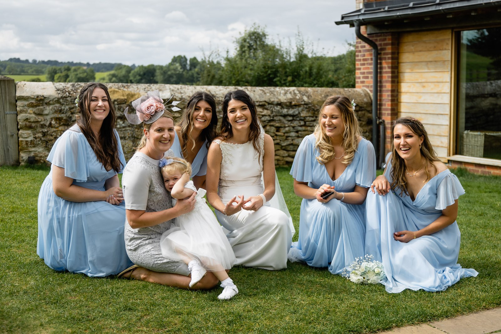 Bridesmaids in powder-blue gowns kneeling on the lawn with a young flower girl