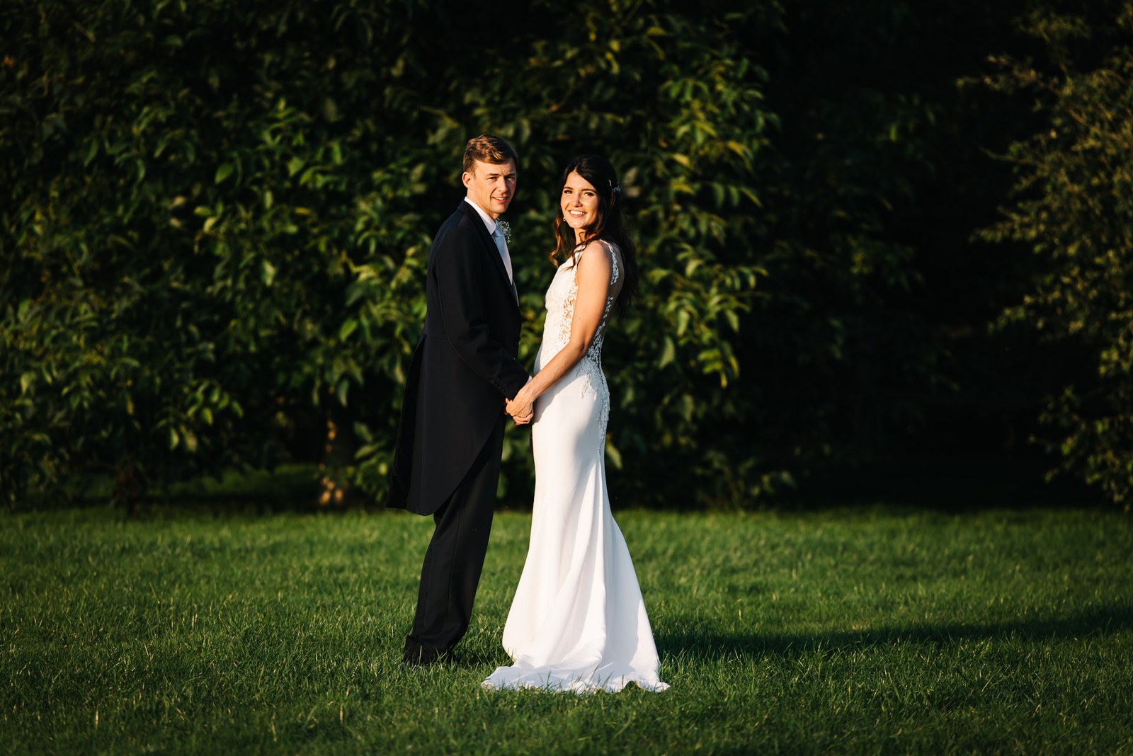 The newlyweds hand in hand on the lawns of Dodford Manor, framed by tall summer trees